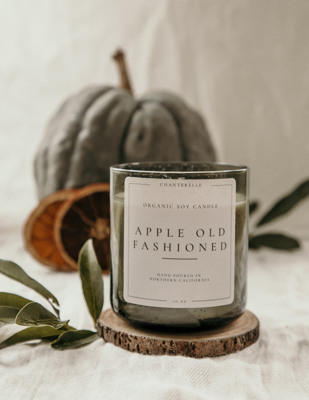 Candle labeled 'Apple Old Fashioned' on a wooden coaster with a pumpkin and leaves in the background.