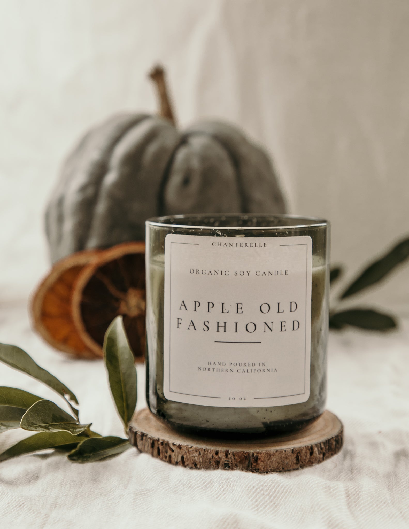 Candle labeled 'Apple Old Fashioned' on a wooden coaster with a pumpkin and leaves in the background.