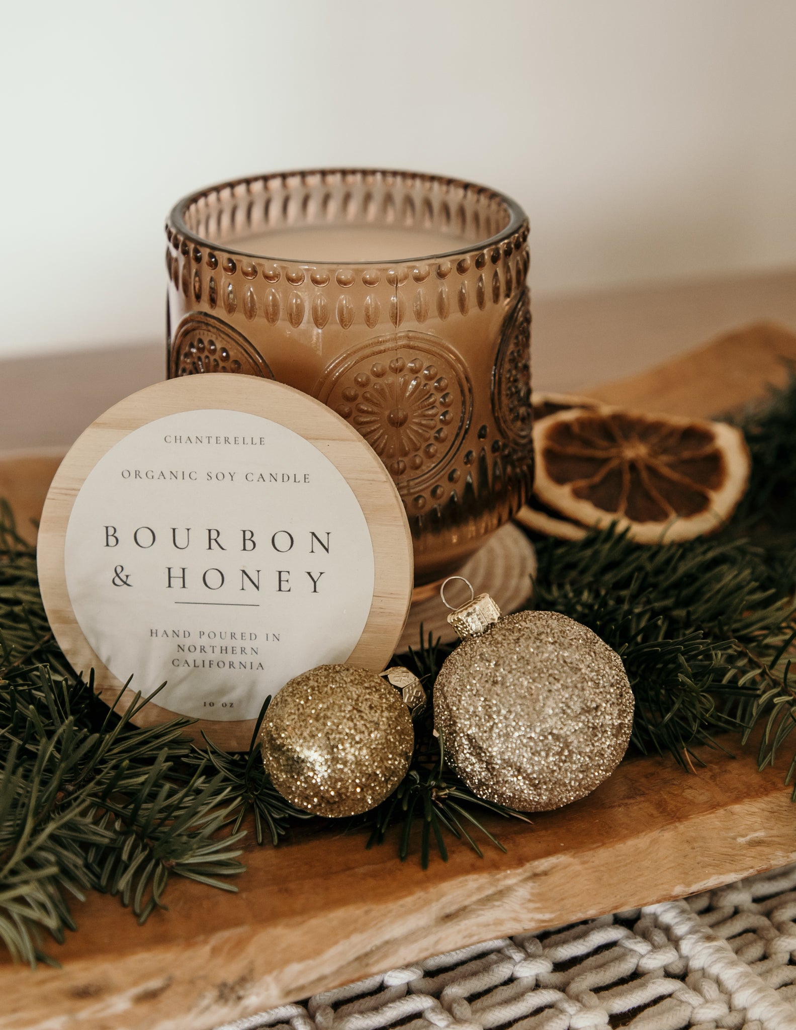 Decorative candle with 'Bourbon & Honey' label, surrounded by gold ornaments and greenery on a wooden surface.
