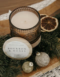 Decorative candle with 'Bourbon & Honey' label on a wooden surface with Christmas decorations.