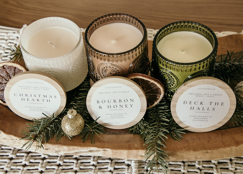 Three candles with decorative labels on a wooden tray with greenery