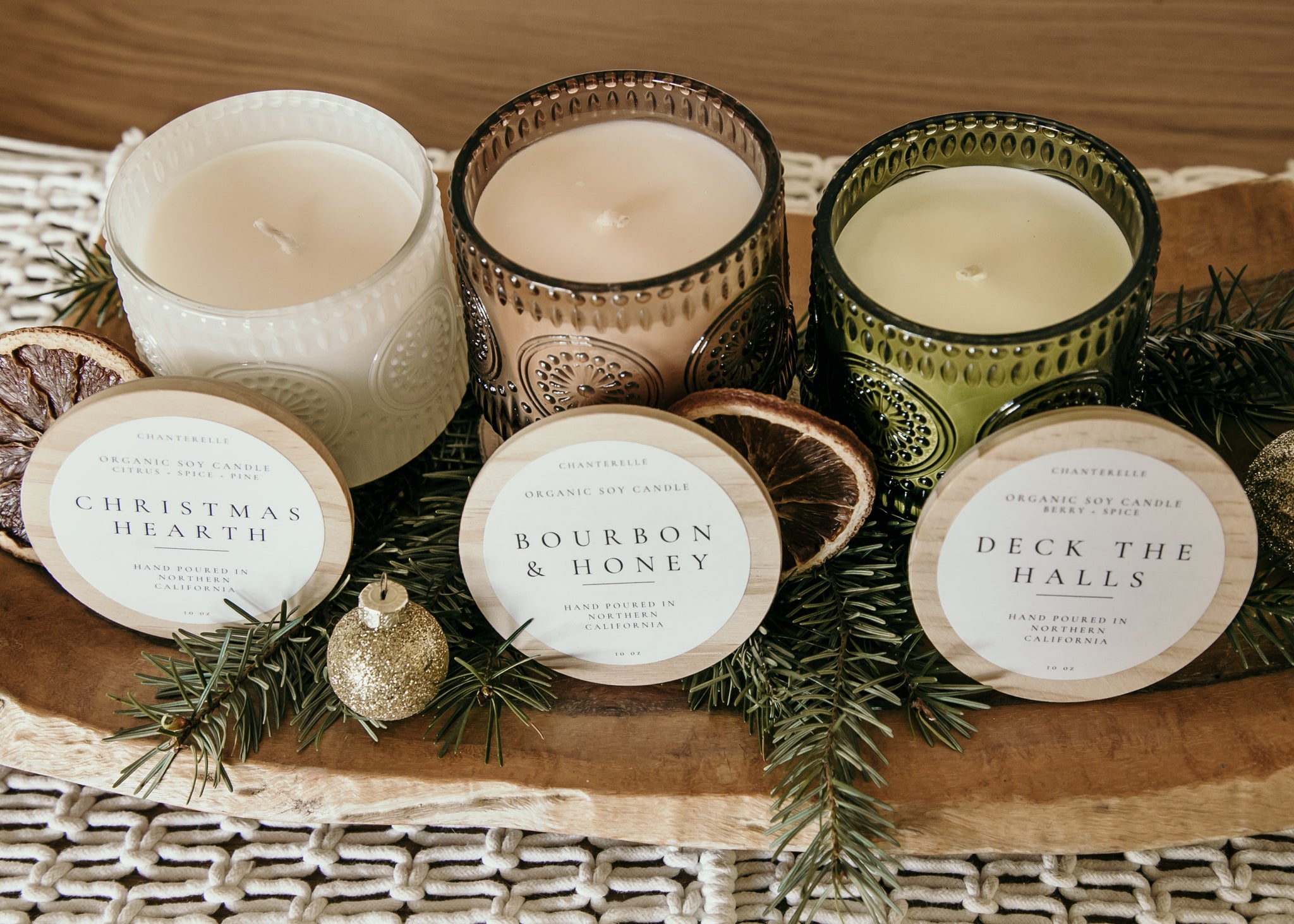Three candles with decorative labels on a wooden tray with greenery