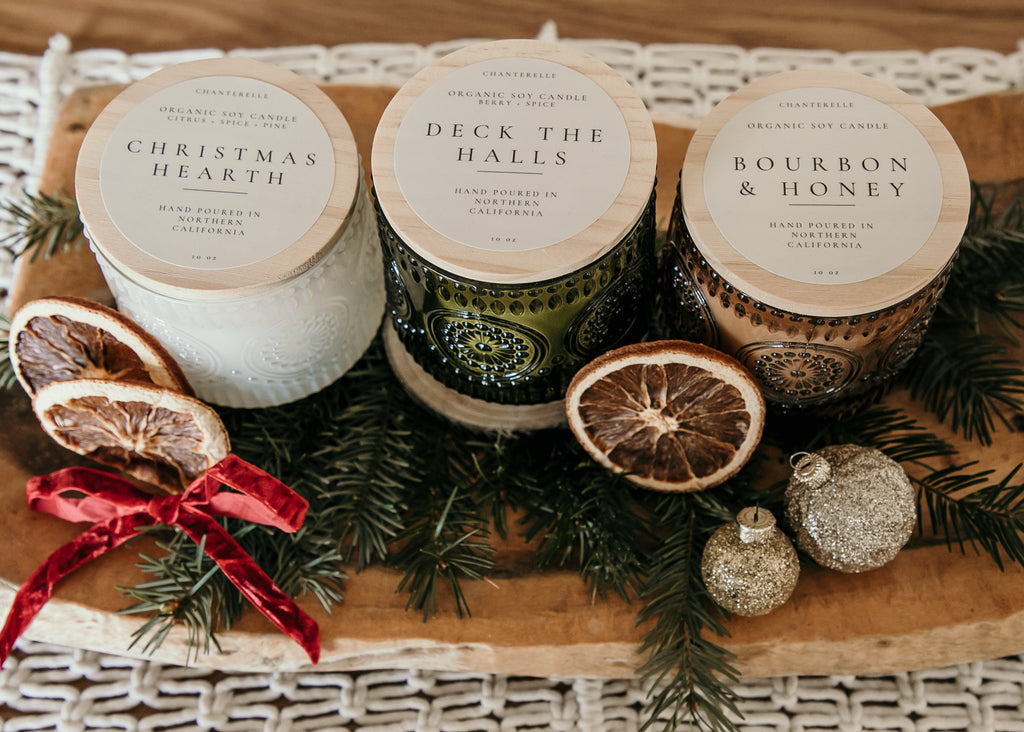 Three candles labeled 'Christmas Hearth', 'Deck the Halls', and 'Bourbon & Honey' on a wooden board with decorative elements.